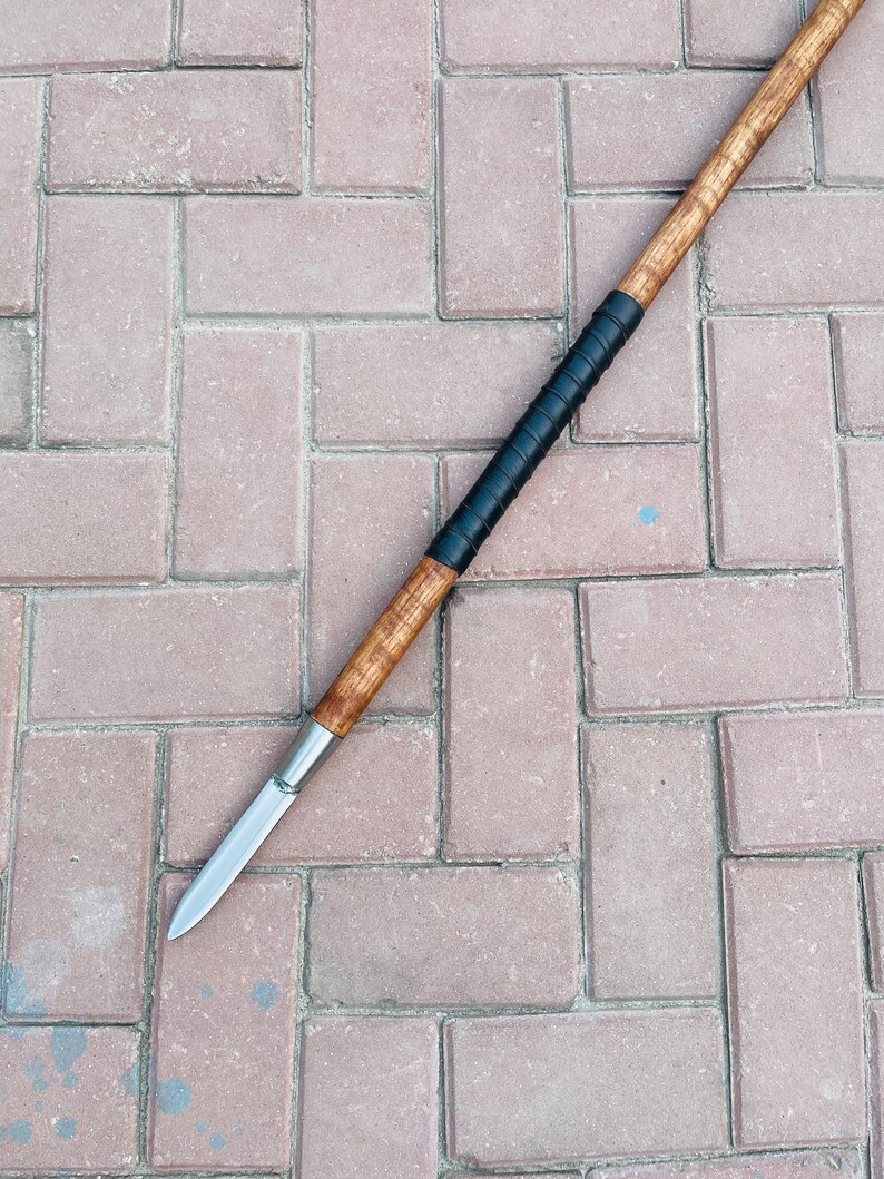 May include: A spear with a wooden shaft and a sharp, silver-colored blade. The shaft has a dark, wrapped grip. The spear is lying on a brick surface.