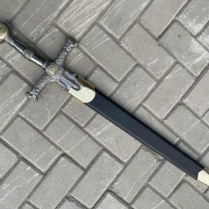 May include: A decorative sword with a silver and gold hilt, featuring a Star of David emblem. The blade is sheathed in a black scabbard with gold accents. The sword rests on a gray brick surface.