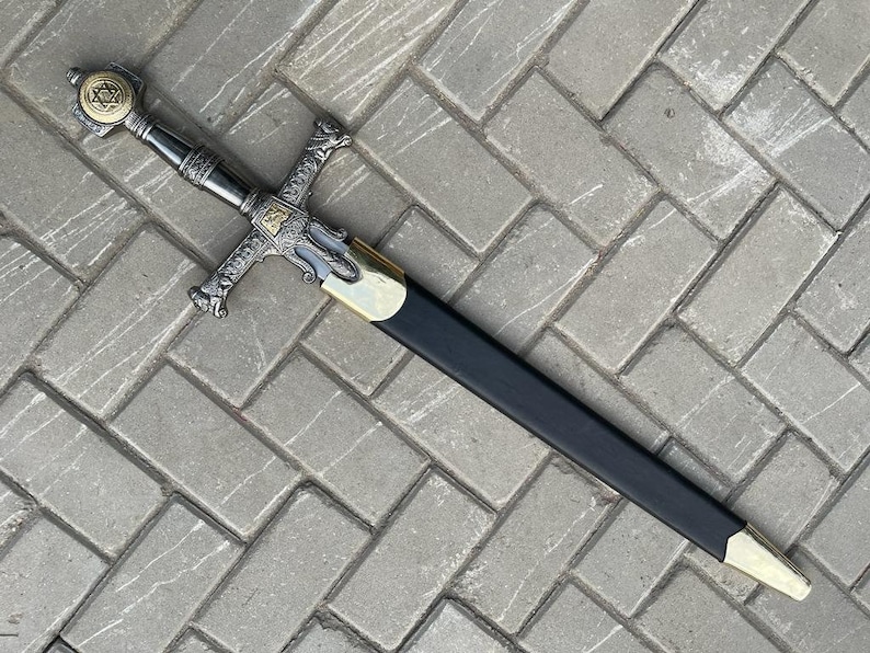 May include: A decorative sword with a silver and gold hilt, featuring a Star of David emblem. The blade is sheathed in a black scabbard with gold accents. The sword rests on a gray brick surface.