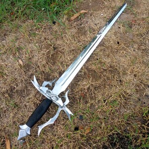May include: A silver-colored sword with an ornate hilt and a black handle lies on the grass. The blade is long and pointed, reflecting the light. The hilt features decorative curves and a detailed design. The sword is a fantasy-themed item.