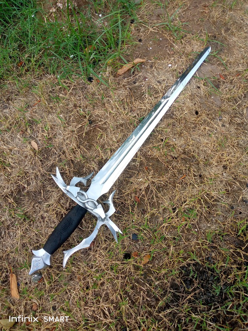May include: A silver-colored sword with an ornate hilt and a black handle lies on the grass. The blade is long and pointed, reflecting the light. The hilt features decorative curves and a detailed design. The sword is a fantasy-themed item.