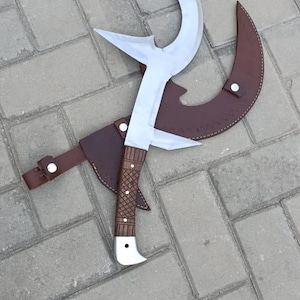 May include: A handmade, curved blade knife with a wooden handle and a brown leather sheath. The blade is silver and has a unique shape. The handle is brown with a textured pattern. The sheath is brown leather with a strap.