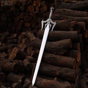 Puede incluir: Una espada plateada con una empu&ntilde;adura ornamentada y una hoja larga y recta. La espada est&aacute; apoyada contra una pila de troncos marrones, creando una est&eacute;tica r&uacute;stica y medieval. El mango presenta detalles intrincados, lo que sugiere una artesan&iacute;a de alta calidad.