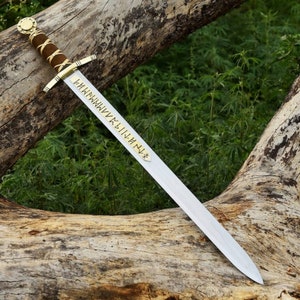 May include: A medieval-style sword with a polished steel blade and a gold-colored crossguard. The handle is wrapped in brown leather. The sword has gold-colored runes etched on the blade. The sword is resting on a weathered log.