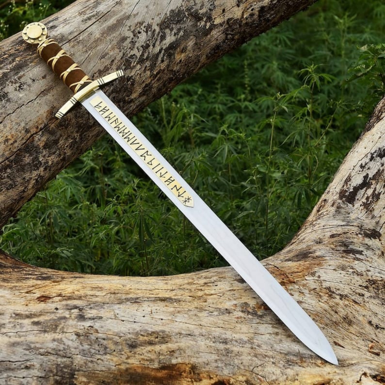 May include: A medieval-style sword with a polished steel blade and a gold-colored crossguard. The handle is wrapped in brown leather. The sword has gold-colored runes etched on the blade. The sword is resting on a weathered log.