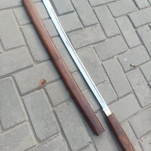 May include: A Japanese-style sword with a polished steel blade and a dark brown wooden scabbard and handle. The blade is straight with a slight curve at the tip. The scabbard is a dark, rich brown color. The handle is also dark brown with a brass accent.
