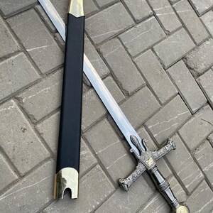 May include: A silver sword with a decorative hilt and a black scabbard with gold accents. The sword is lying on a gray brick surface. The hilt features intricate designs and a gold emblem.