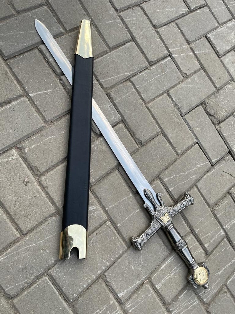 May include: A silver sword with a decorative hilt and a black scabbard with gold accents. The sword is lying on a gray brick surface. The hilt features intricate designs and a gold emblem.