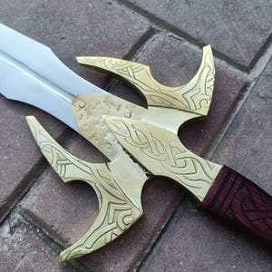May include: A close-up of a decorative sword with a silver blade and a brass hilt. The hilt features intricate Celtic knot designs and a dark red wooden handle. The sword is lying on a brick surface.