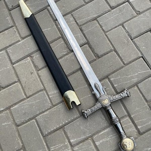 May include: A decorative sword with a silver blade and ornate hilt. The hilt features gold accents and a detailed design. The black scabbard has gold-colored metal accents. The sword is displayed on a gray brick surface.