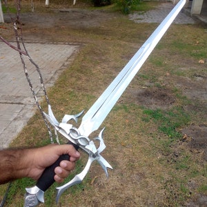 May include: A silver-colored sword with an ornate hilt and a long, double-edged blade. The hilt features intricate designs and a black grip. The sword is held against a backdrop of green grass and a blurred background.