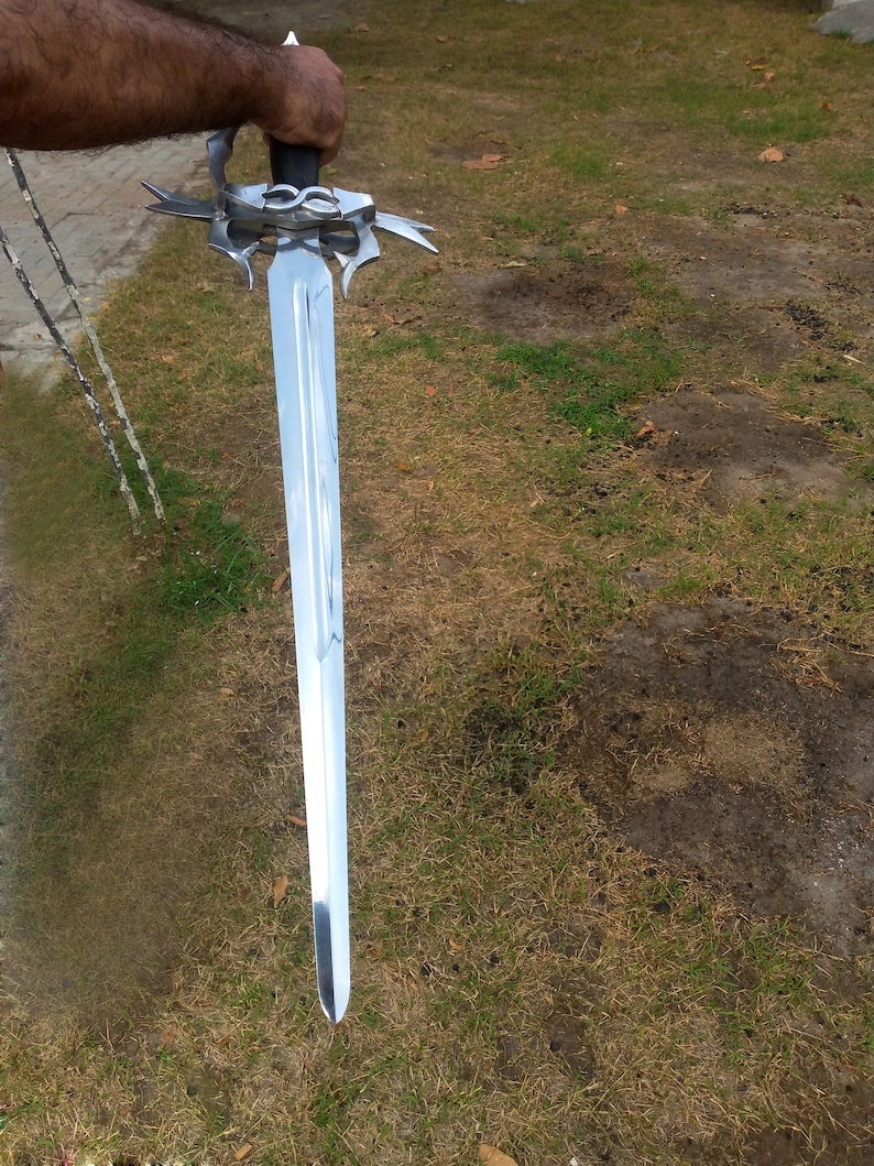 May include: A long, silver sword with a decorative hilt and a black handle. The blade is highly polished, reflecting the surrounding environment. The sword is held against a backdrop of green grass.