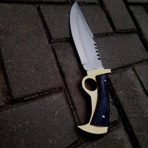 May include: A large, stainless steel knife with a polished blade and a brass and dark blue handle. The handle has a unique, curved design. The knife is designed for utility and is suitable for various tasks.