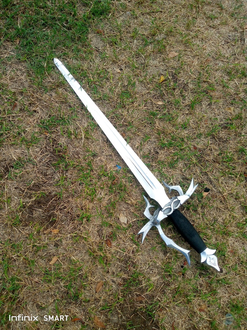 May include: A silver-colored sword with a black handle lies on a bed of grass. The sword has a long, pointed blade and an ornate hilt with decorative elements. The handle appears to be wrapped in black material.