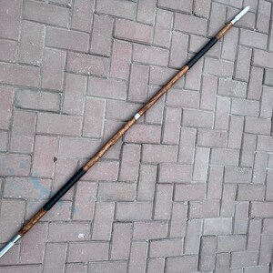 May include: A spear with a wooden shaft and a metal spearhead. The shaft is brown with black wrapped sections near the ends. The spearhead is silver and pointed. The spear is lying on a brick surface.