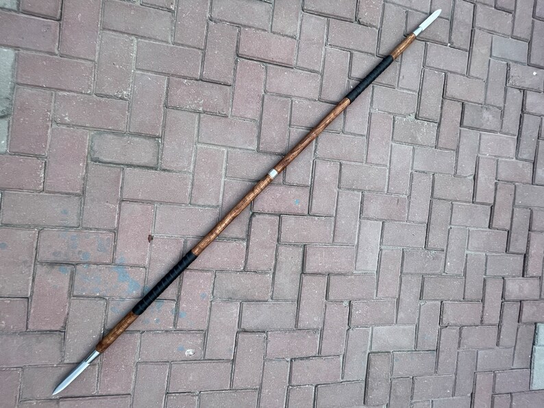 May include: A spear with a wooden shaft and a metal spearhead. The shaft is brown with black wrapped sections near the ends. The spearhead is silver and pointed. The spear is lying on a brick surface.