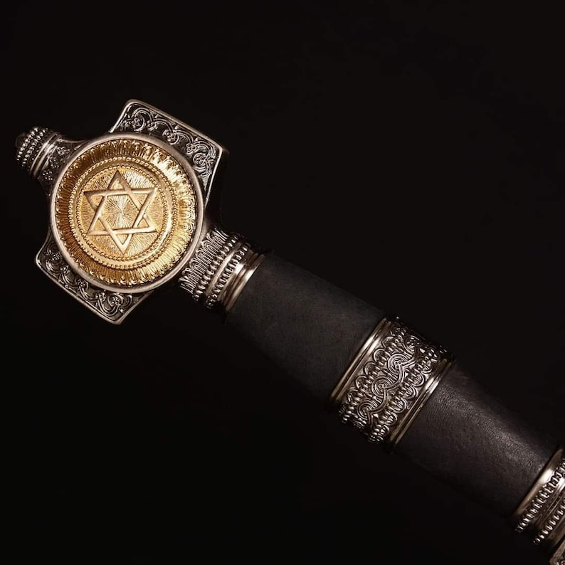 May include: Close-up of a decorative sword hilt with a gold Star of David emblem. The hilt features ornate silver detailing and a black leather grip, set against a dark background.