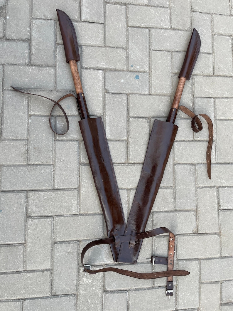 May include: Two brown leather sheathed knives with wooden handles are displayed on a gray brick surface. The knives are positioned in a V-shape, with leather straps and a belt. The sheaths are dark brown.