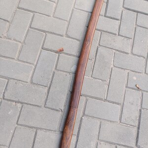May include: A long, wooden scabbard with a rich, dark brown finish. The scabbard is straight with a slight curve and a visible seam near the bottom. The wood grain is prominent, adding texture to the surface.