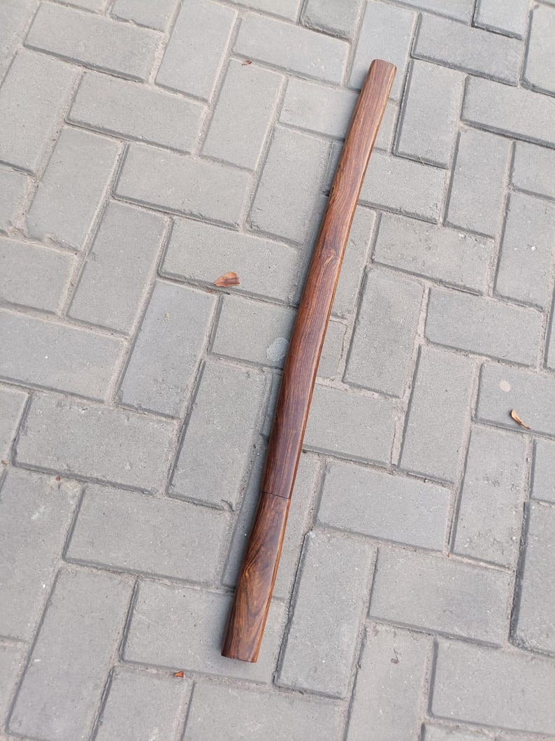 May include: A long, wooden scabbard with a rich, dark brown finish. The scabbard is straight with a slight curve and a visible seam near the bottom. The wood grain is prominent, adding texture to the surface.