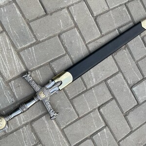May include: A decorative sword with a black scabbard and ornate silver and gold hilt. The sword features a cross-shaped guard and a detailed handle with a circular emblem. The blade is not visible.
