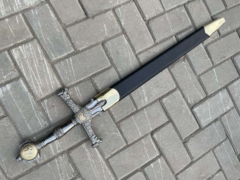May include: A decorative sword with a black scabbard and ornate silver and gold hilt. The sword features a cross-shaped guard and a detailed handle with a circular emblem. The blade is not visible.