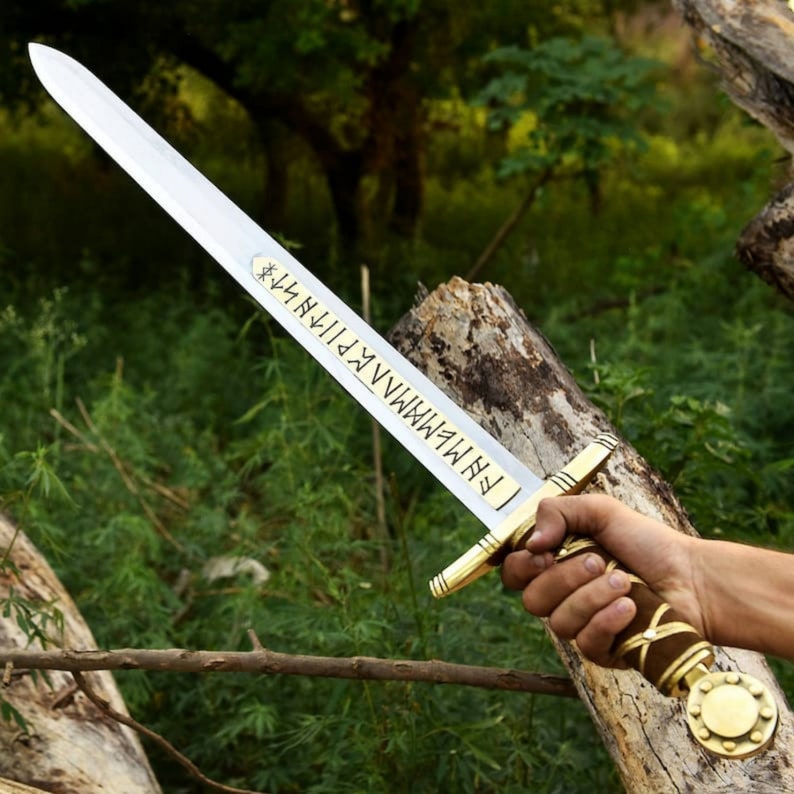 May include: A long, silver sword with a gold hilt and a brown leather-wrapped handle. The blade has etched runes. The sword is held against a backdrop of green foliage and tree trunks.