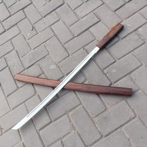 May include: A Japanese-style sword with a polished steel blade and a dark wood handle. The sword is lying on a gray brick surface, with a matching wooden scabbard nearby. The blade is approximately 24 inches long.