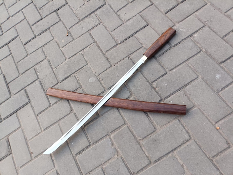 May include: A Japanese-style sword with a polished steel blade and a dark wood handle. The sword is lying on a gray brick surface, with a matching wooden scabbard nearby. The blade is approximately 24 inches long.