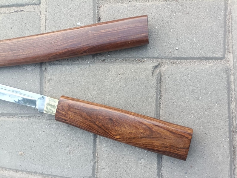 May include: A close-up of a sword with a polished steel blade and a wooden handle. The handle is a rich brown color with a brass accent. The scabbard is also made of wood, matching the handle's color.