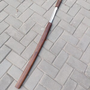 May include: A long, slender, wooden object with a dark brown finish. The object has a rectangular shape with a silver metal insert. The item is lying on a gray brick surface.