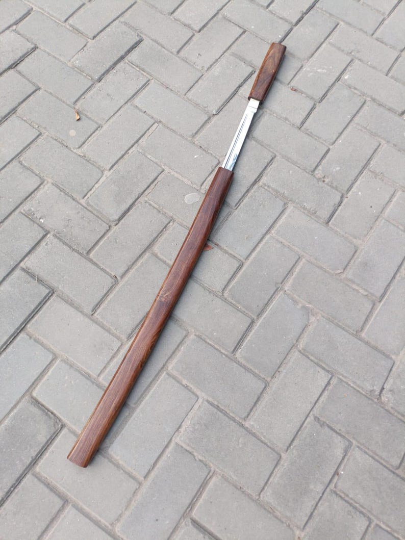 May include: A long, slender, wooden object with a dark brown finish. The object has a rectangular shape with a silver metal insert. The item is lying on a gray brick surface.