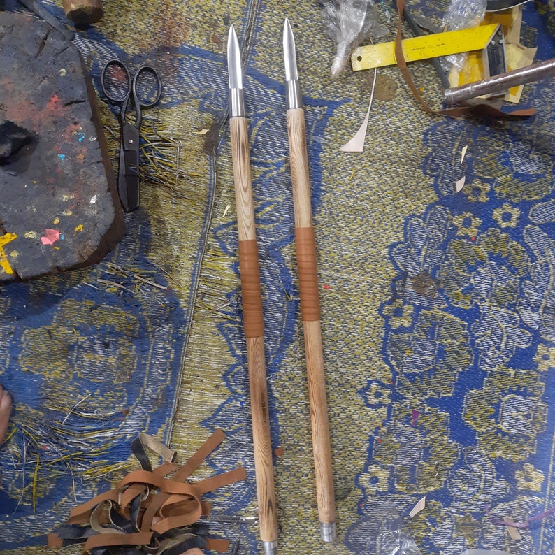 May include: Two spears with wooden shafts and metal spearheads. The shafts have a light brown wood grain pattern and are wrapped with brown leather near the center. The spears are lying on a blue and yellow patterned surface.