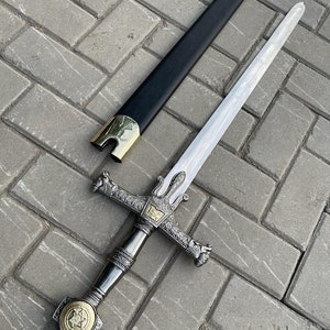 May include: A decorative sword with a silver blade and ornate hilt. The hilt features intricate detailing and a gold-colored emblem. The black scabbard has gold-colored accents. The sword is displayed on a gray brick surface.