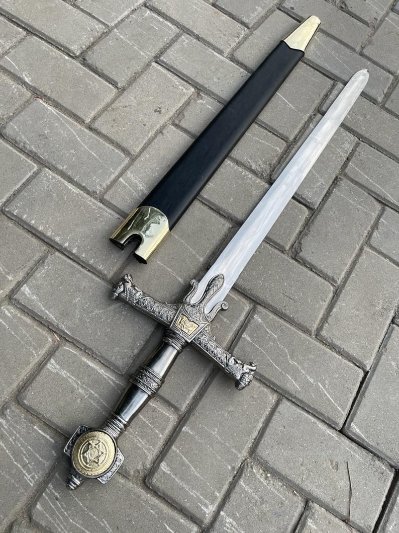 May include: A decorative sword with a silver blade and ornate hilt. The hilt features intricate detailing and a gold-colored emblem. The black scabbard has gold-colored accents. The sword is displayed on a gray brick surface.