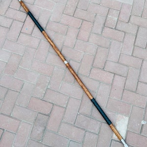 May include: A double-edged spear with a wooden shaft, metal spearheads, and black wrapped grip. The spear is approximately 72 inches long, lying on a brick surface.