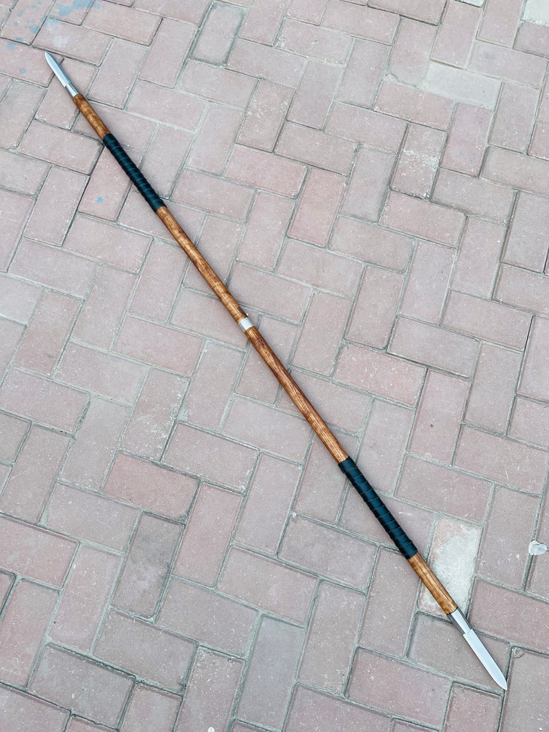 May include: A double-edged spear with a wooden shaft, metal spearheads, and black wrapped grip. The spear is approximately 72 inches long, lying on a brick surface.
