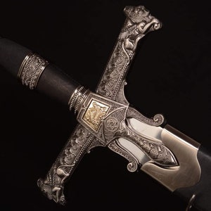 May include: An ornate sword with a silver-colored hilt and a black handle. The hilt features intricate engravings, including floral patterns and a gold-colored emblem. The blade is partially visible, and the overall design suggests a historical or ceremonial purpose.