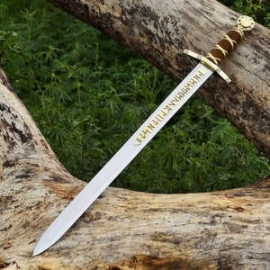 May include: A long, silver sword with a brown leather-wrapped handle and gold accents. The blade features engraved text and rests on a weathered log against a backdrop of green foliage. The sword's design suggests a historical or fantasy theme.