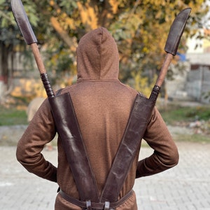 May include: A brown leather back sheath designed to hold two large knives. The sheath is worn over the shoulders and around the waist. The knives have wooden handles and leather sheaths. The person is wearing a brown hooded sweatshirt.