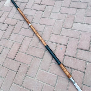 May include: A spear with a wooden shaft, black grip, and a silver blade. The spear is approximately 70 inches long. The shaft has a light brown wood grain pattern. The grip is wrapped in black material. The blade is silver and pointed.