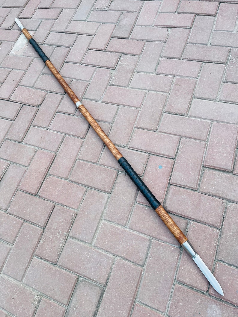 May include: A spear with a wooden shaft, black grip, and a silver blade. The spear is approximately 70 inches long. The shaft has a light brown wood grain pattern. The grip is wrapped in black material. The blade is silver and pointed.
