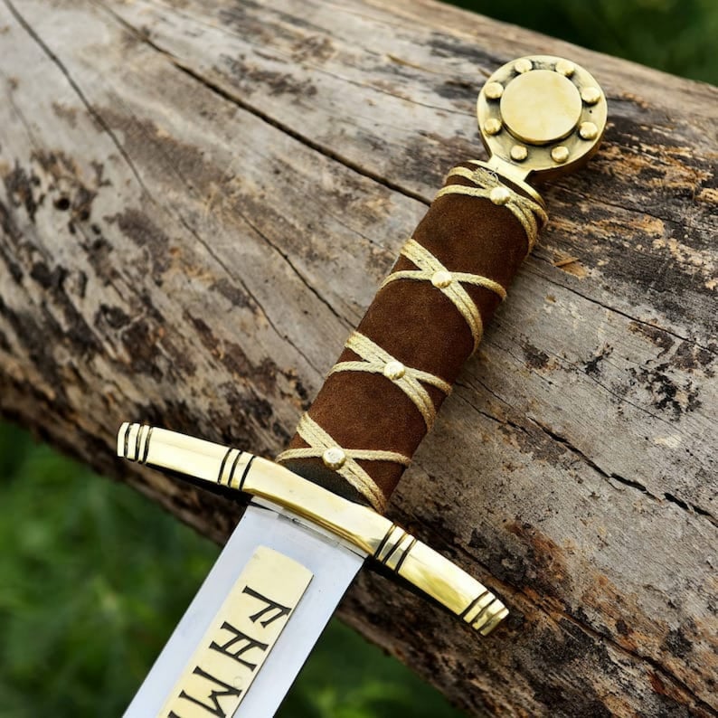 May include: A close-up of a decorative sword with a brown suede handle wrapped with gold-colored cord. The sword features a gold-colored hilt and a blade with etched text. The sword rests on a weathered wooden surface.