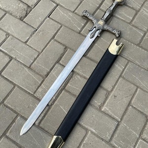 May include: A silver and gold decorative sword with a black sheath. The sword features an ornate hilt and a long, straight blade. The sheath has gold-colored accents on the tip and opening.