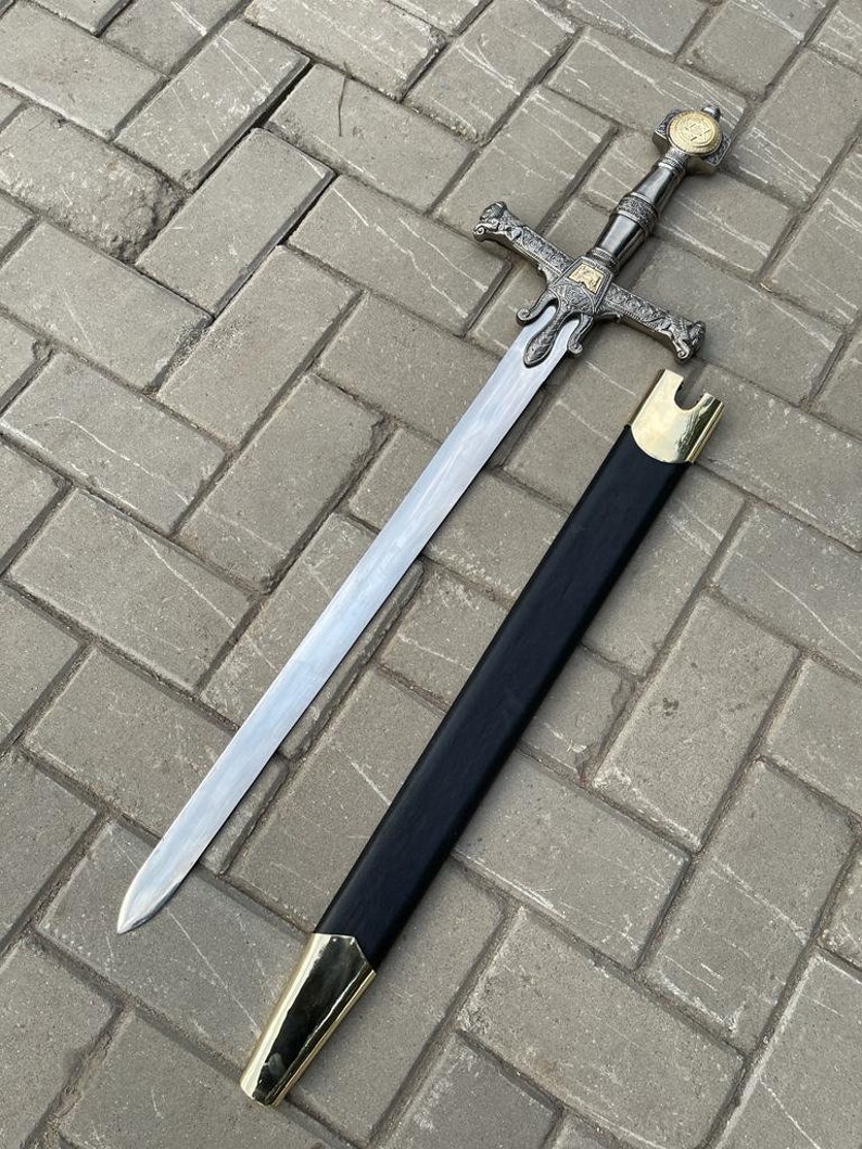 May include: A silver and gold decorative sword with a black sheath. The sword features an ornate hilt and a long, straight blade. The sheath has gold-colored accents on the tip and opening.