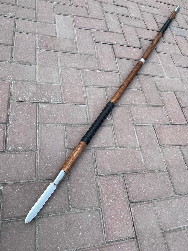 May include: A spear with a wooden shaft and a silver blade. The shaft is a dark brown color with a black grip near the center. The spear is lying on a brick surface.