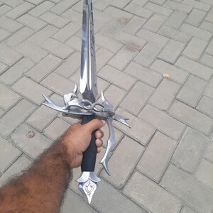 May include: A polished steel sword with an ornate hilt and a black grip. The blade is long and double-edged. The hilt features decorative elements and a stylized floral design at the pommel. The sword is held in a hand.