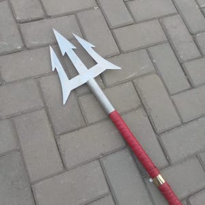 May include: A silver trident with three prongs, a gray shaft, and a red wrapped handle. The handle is accented with a gold band. The trident is lying on a gray brick surface.
