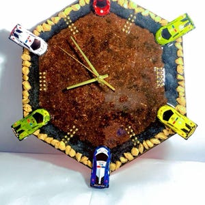 May include: A unique, hexagonal clock with a brown center and a black border. The clock is decorated with small toy cars in red, white, blue, and yellow. The clock hands are green, and the border is lined with small, yellow stones.