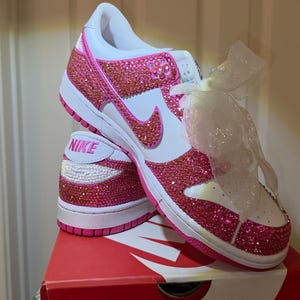 May include: Customised Nike Dunk Low trainers, featuring pink rhinestones. The shoes have a white base with pink detailing, including the Nike swoosh and sole. The trainers are displayed on a red Nike box.